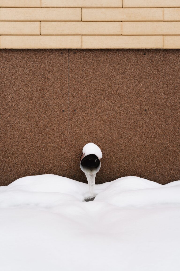 Frozen pipe with frost and icicles against a snow-covered building wall in winter