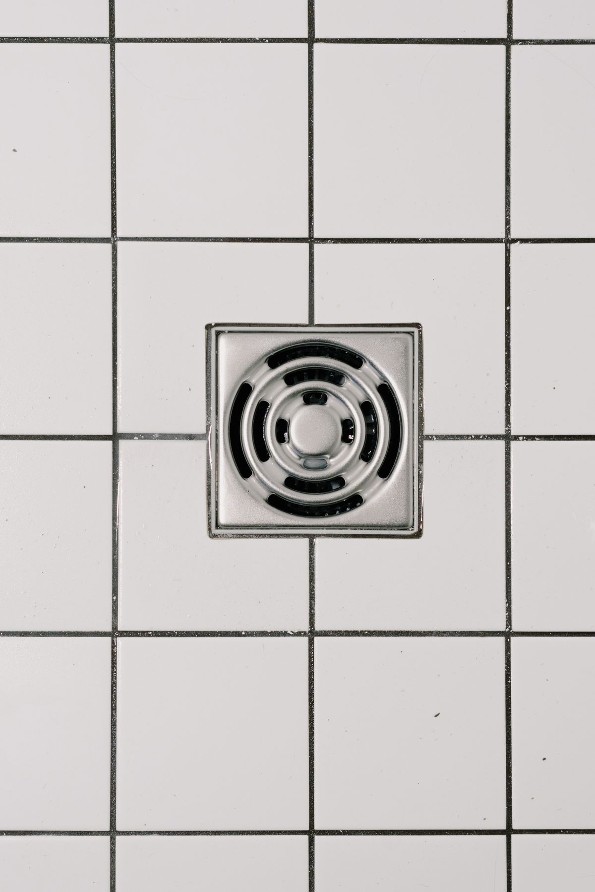 Square stainless-steel shower drain cover set into white bathroom floor tiles